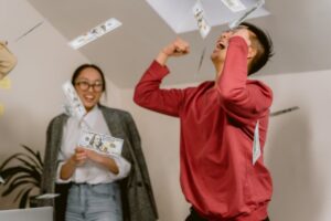 Two young adults in a room celebrating with falling dollar bills, expressing excitement and joy.